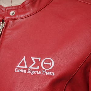 May include: A close-up of a red leather jacket with a silver zip and a snap closure at the collar. The jacket features the embroidered Greek letters Delta Sigma Theta in white.