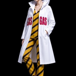 Op de afbeelding: Een witte lange jas met capuchon met het woord GAS in opvallende roze letters aan beide kanten van de borst, gedragen over een mouwloze top en broek met gele en zwarte diagonale strepen, gecombineerd met witte sneakers.