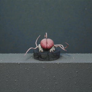 May include: A detailed sculpture of a brain-like creature with pink and red tones, featuring multiple tendrils. The creature rests on a dark, textured platform, set against a muted gray backdrop. The sculpture has a unique, otherworldly appearance.