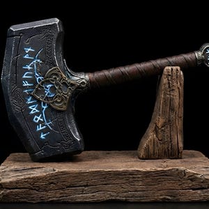 May include: A detailed replica of Thor's hammer, Mjolnir, resting on a wooden stand. The hammer features a dark gray head with blue glowing runes and intricate metalwork. The handle is wrapped in brown leather. The stand is made of rough-hewn wood, set against a black background.