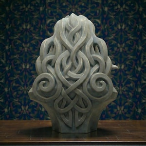 May include: A grey stone-like sculpture featuring an intricate, raised relief of interconnected, flowing knotwork patterns. The design consists of symmetrical, swirling loops and interlaced bands that create a dense, textured surface.