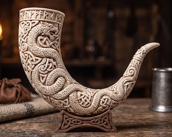 May include: A cream-colored drinking horn featuring intricate relief carvings of a coiled serpent and Celtic knotwork patterns. The rim is decorated with engraved runic characters. The horn rests on a dark brown wooden stand with carved details.