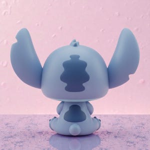 May include: A light blue figurine of the character Stitch from Lilo & Stitch, viewed from the back. The figurine has large ears, a distinctive back pattern, and is sitting down. The background is a light pink color.