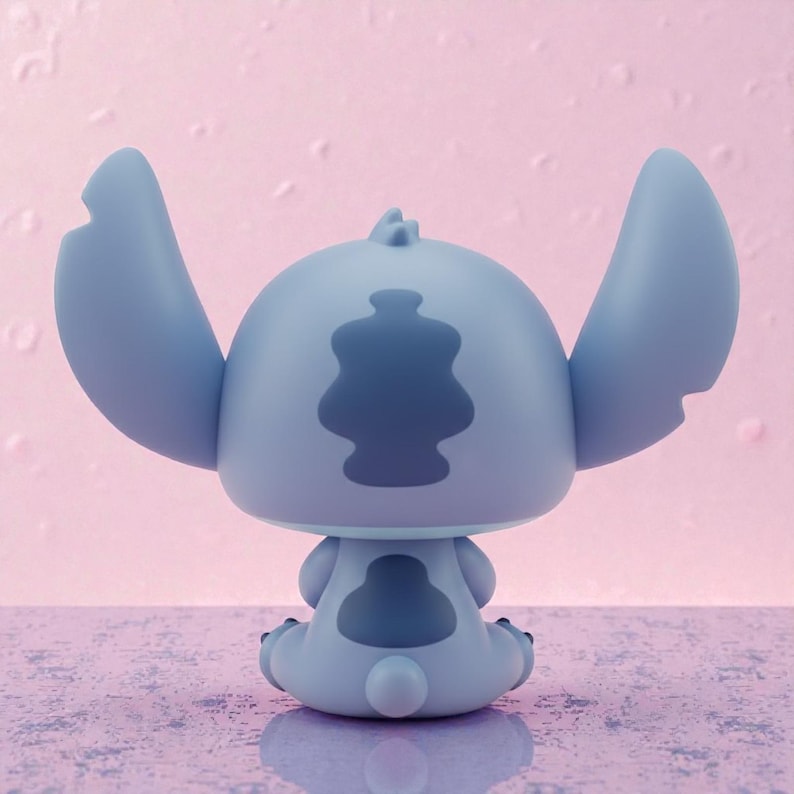 May include: A light blue figurine of the character Stitch from Lilo & Stitch, viewed from the back. The figurine has large ears, a distinctive back pattern, and is sitting down. The background is a light pink color.