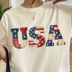 May include: Off-white t-shirt with the word "USA" in red, white, and blue, resembling the American flag. The letters are embroidered with a vintage, slightly distressed look. The shirt has short sleeves and a crew neck.