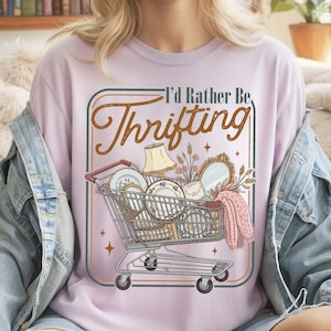 A vintage-inspired digital PNG design featuring a retro shopping cart filled with antique treasures like porcelain plates, a gold mirror, and a lamp. Above the cart, the text 'I'd Rather Be Thrifting' is written in a stylish brown script. The design has a muted, boho color palette and a transparent background, perfect for t-shirt sublimation and crafting.