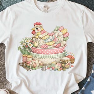 May include: White t-shirt featuring a colorful illustration of a patchwork hen sitting in a bowl. The design includes sewing notions, flowers, and a watering can. The hen is adorned with a bow and a tag.