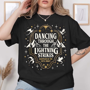 Puede incluir: Camiseta negra con un diseño gráfico dorado y blanco. El diseño presenta el texto "Dancing Through The Lightning Strikes" con ilustraciones de montañas y dragones. También se incluye la frase "Sleepless in the Onyx Night".