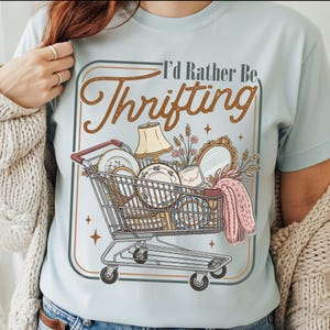 PNG digital design featuring a vintage-style illustration of a shopping cart filled with thrifted home decor items and the phrase “I’d Rather Be Thrifting” in retro typography, shown on a t-shirt mockup.
