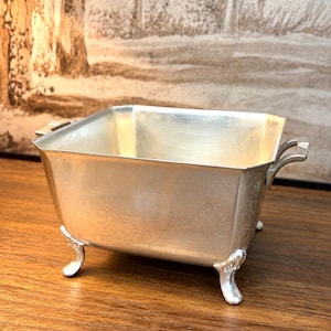 May include: A rectangular silver-toned metal bowl with a smooth interior and a subtle textured exterior. The vessel features two small handles on opposite sides and is supported by four ornate, curved feet at each corner of the base.