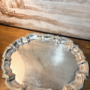 May include: A round silver-toned serving tray featuring a scalloped, raised edge and an intricate, engraved floral pattern across the center surface. The metal tray has a polished, reflective finish and a distinctively shaped, wavy rim.