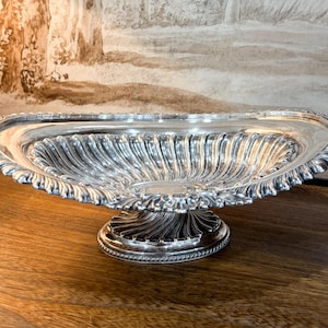 May include: An oval silver bowl with a pedestal base, featuring a reflective metallic finish and intricate fluted detailing along the interior basin and the exterior stem. The rim is decorated with a scalloped, embossed edge pattern.