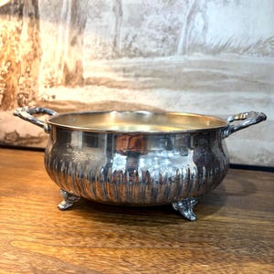 May include: A silver-toned metal bowl with a polished finish, featuring a rounded body with vertical fluted detailing, two ornate side handles, and three small decorative feet. The interior of the bowl has a smooth, reflective surface.