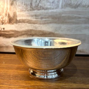 May include: A polished silver bowl with a smooth, reflective surface and a rounded body. The bowl features a wide, circular opening and rests on a sturdy, elevated pedestal base with a tiered design, showcasing a clean and metallic finish.