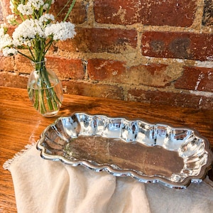 May include: A silver-colored, rectangular serving tray with a scalloped edge sits on a cream-colored linen cloth. A small glass vase with white flowers is to the left. The background is a rustic brick wall.