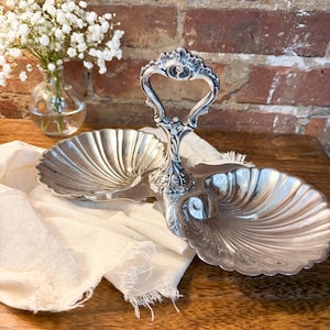May include: A silver-colored, two-tiered serving dish with a decorative handle. The dish has a scalloped design and sits on a wooden surface. A small vase of white flowers is in the background.