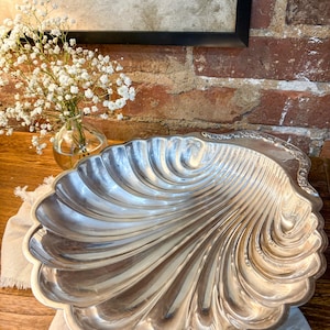 May include: A large, silver-colored serving dish in the shape of a seashell. The dish has a scalloped edge and a textured, ridged surface. The dish is sitting on a white cloth, with a vase of white flowers in the background.
