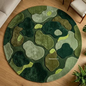 Op de afbeelding: Een ronde, groene vloerkleed met een abstract patroon van verschillende groentinten, van licht limoengroen tot diep bosgroen. Het kleed ligt op een houten vloer, met een bank, stoel en planten op de achtergrond. Het patroon van het kleed lijkt op een camouflagepatroon.
