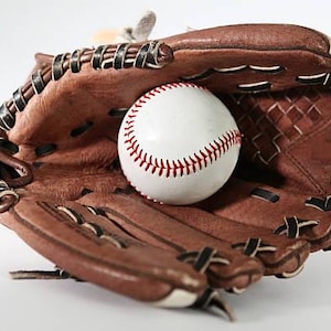 May include: A brown leather baseball glove with a white baseball inside. The baseball has red stitching. The glove is open, ready to catch the ball. The image is set against a white background.