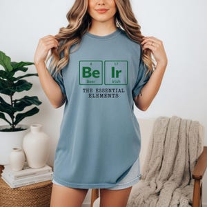 May include: A light blue t-shirt with a green graphic design. The design features the words "Be Ir" in a periodic table style, with "Beer" and "Irish" below. The text "THE ESSENTIAL ELEMENTS" is below the graphic.