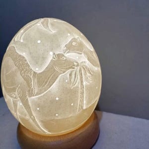 Pode incluir: Uma escultura em forma de ovo de cor creme com entalhes detalhados de dois camelos perto de uma palmeira. A superfície tem uma aparência texturizada e salpicada, montada sobre uma base circular lisa de madeira castanho-escura.