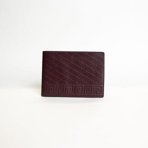 Może przedstawiać: Bordowy skórzany portfel z geometrycznym wzorem i greckim wzorem klucza. Portfel jest prostokątny i wydaje się nowy.