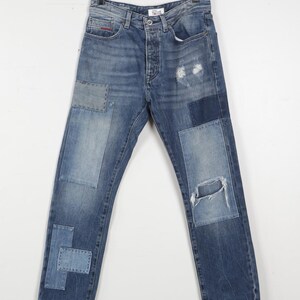 Könnte beinhalten: Blaue Jeans mit mehreren Aufnähern in verschiedenen Blautönen. Die Jeans haben einen Used-Look mit einem zerrissenen Knie. Die Jeans hängen an einem schwarzen Kleiderbügel.