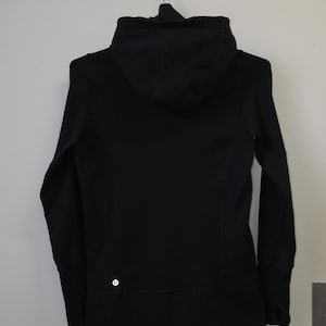 May include: A black hooded jacket with a zipper closure, long sleeves, and a small logo on the lower left side. The jacket is hanging on a white wall. The jacket is made of a soft, flexible material.