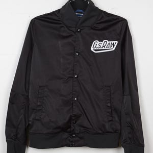 May include: Black satin bomber jacket with a ribbed collar, cuffs, and hem. The jacket features a button-up front and a white logo that reads "G.S. RAW" on the chest. The jacket is hanging on a metal hook.