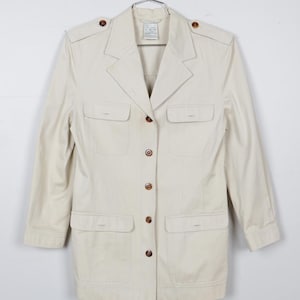 May include: Cream-colored jacket with four front pockets and brown buttons. The jacket has a collared neckline and shoulder epaulets. It is hanging on a metal hanger against a white background.