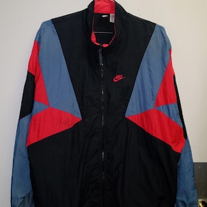 Puede incluir: Chaqueta cortavientos Nike vintage en negro, rojo y azul. La chaqueta tiene cremallera completa, cuello alto y el logo de Nike bordado en rojo en el pecho. Las mangas son azules y la parte delantera tiene paneles rojos y azules.