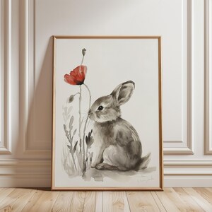 Puede incluir: Impresión en acuarela enmarcada de un conejo gris y una flor de amapola roja. La obra presenta una ilustración detallada del conejo con largas orejas y una cola esponjosa, sobre un fondo blanco. La amapola proporciona un toque de color vibrante.