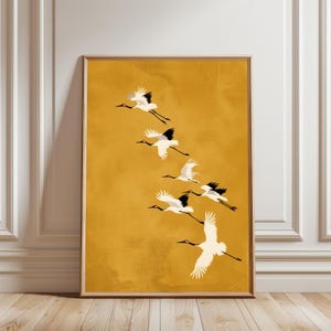 Puede incluir: Una impresión artística enmarcada que presenta una bandada de grullas blancas y negras en vuelo sobre un fondo amarillo dorado. Las aves están representadas con las alas extendidas, creando una sensación de movimiento y gracia. La obra de arte está en un marco de madera.