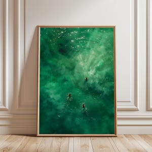 Op de afbeelding: Een ingelijste kunstdruk met een luchtfoto van drie mensen die zwemmen in heldergroen water. Het kunstwerk legt het samenspel van licht en schaduw vast, waardoor een gevoel van diepte en rust ontstaat. De print is ingelijst in een natuurlijke houten lijst.