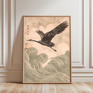 Puede incluir: Lámina artística enmarcada que representa una grulla japonesa en vuelo sobre olas estilizadas. La grulla está representada en tonos grises con la cabeza roja, sobre un fondo beige y nublado. La obra está enmarcada en un marco de madera marrón claro.