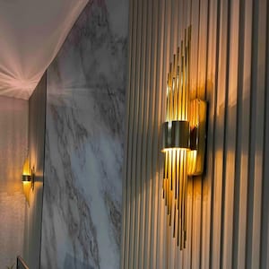 Puede incluir: Apliques de pared dorados con un diseño vertical, montados en una pared clara con líneas verticales. Los apliques tienen una base cilíndrica y una serie de varillas verticales que crean una forma de abanico. La pared detrás de los apliques tiene un patrón similar al mármol.