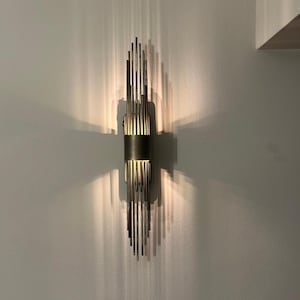 May include: A contemporary wall sconce featuring a black cylindrical band and vertical metal rods. The sconce emits warm light, creating linear shadows on the grey wall. The fixture is mounted on a neutral-toned wall.