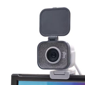 May include: A gray and white webcam with a built-in privacy cover. The webcam has a square design with a lens and the "logi" logo. It is mounted on a computer monitor with a white cable.