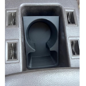 May include: A black, 3D-printed cup holder designed for a vehicle's centre console. The cup holder has a circular opening and a rectangular base, with a textured surface. The surrounding console is brown.