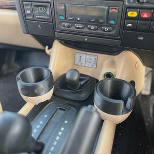 Puede incluir: Vista interior del salpicadero y la consola central de un vehículo. El salpicadero cuenta con un reloj digital y controles de radio. La consola incluye una palanca de cambios, portavasos y una toma de corriente. La combinación de colores es negro y beige.