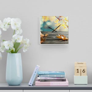 May include: Square wall clock with a nature-themed image. The clock face features a wooden surface with fallen leaves and a blurred background of yellow and green foliage. Black clock hands indicate the time.