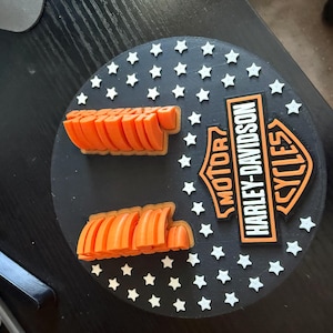 May include: A black circular decorative piece with white star accents and the Harley-Davidson Cycles logo in orange and white. Two orange, stylized, arched shapes are positioned above and below the logo.