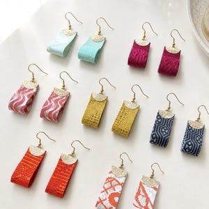 Puede incluir: Seis pares de pendientes colgantes con cintas de tela plegadas en varios colores, incluyendo menta, magenta, rosa, amarillo, azul marino, naranja y blanco con patrones naranjas. Cada pendiente tiene un remate semicircular dorado y gancho.