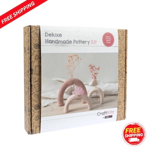 May include: A 'Deluxe Handmade Pottery Kit' box featuring three arch-shaped vases in shades of pink and white, each holding dried flowers. The box has a brown patterned design and the text "Deluxe Handmade Pottery Kit".