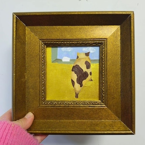 Op de afbeelding: Een ingelijst schilderij met een kat met bruine vlekken, die uit een raam kijkt. De achtergrond toont een gele muur, een blauwe lucht met wolken en een wit kozijn. De lijst is goudkleurig.