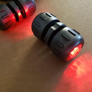 Puede incluir: Primer plano de varios objetos cilíndricos grises con un diseño segmentado y una luz roja que emana de un extremo. Los objetos están dispuestos sobre una superficie de tono neutro, creando un contraste vibrante.