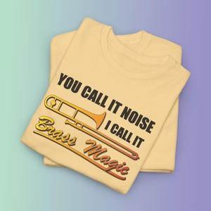 Op de afbeelding: Twee lichtgele T-shirts op elkaar. Het bovenste shirt heeft een trombone-afbeelding met de tekst "YOU CALL IT NOISE I CALL IT BRASS MAGIC" in zwart, geel en oranje. De shirts zijn op een verloopachtergrond gevouwen.