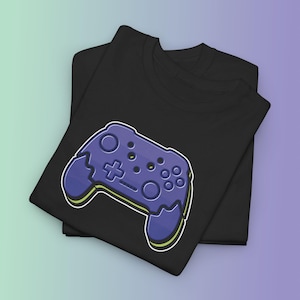 May include: Two black t-shirts are stacked, with the top shirt featuring a large, stylized purple video game controller graphic. The controller has a white outline and yellow accents. The background is a gradient of green and purple.