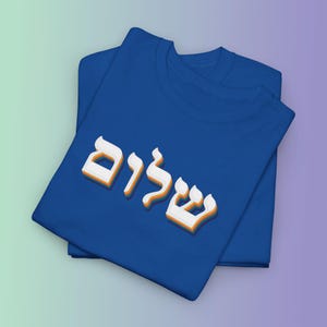 May include: Two folded royal blue t-shirts. The top shirt features the Hebrew word "Shalom" in white, outlined in orange. The shirts are displayed on a gradient background of purple and green.