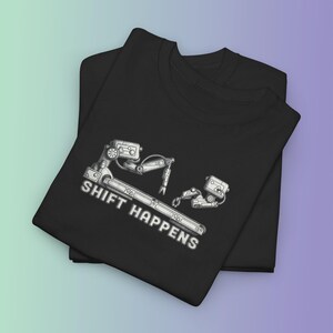 May include: A black t-shirt featuring a white graphic illustration of two mechanical robotic arms positioned above a long metal bar. The text SHIFT HAPPENS is printed in white capital letters below the robotic arms and the metal bar.
